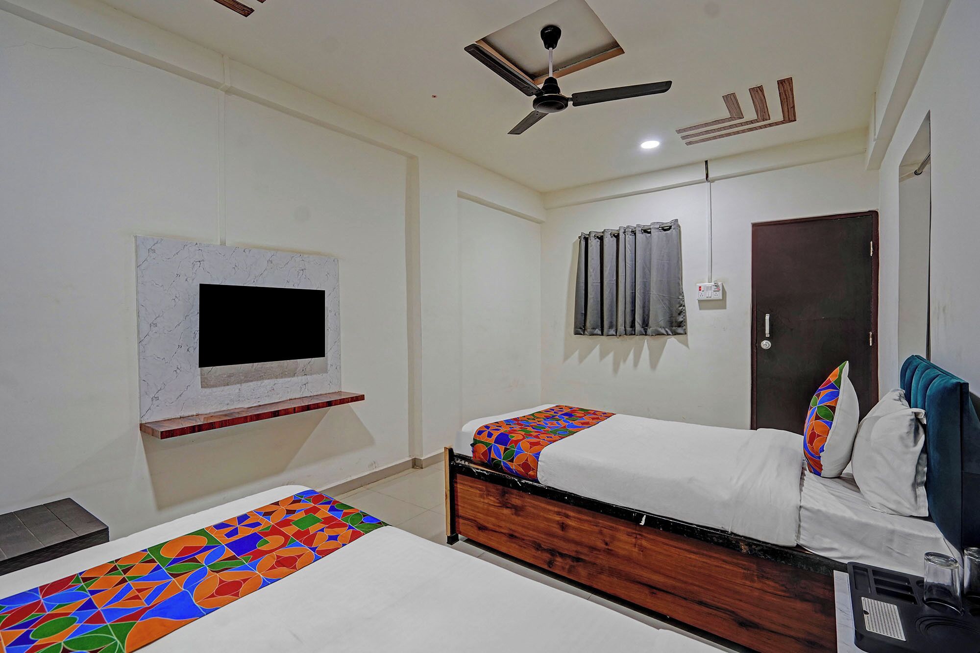 Quarto de hotel com duas camas, um ventilador de teto, uma televisão fixada na parede e uma porta com uma toalha pendurada.