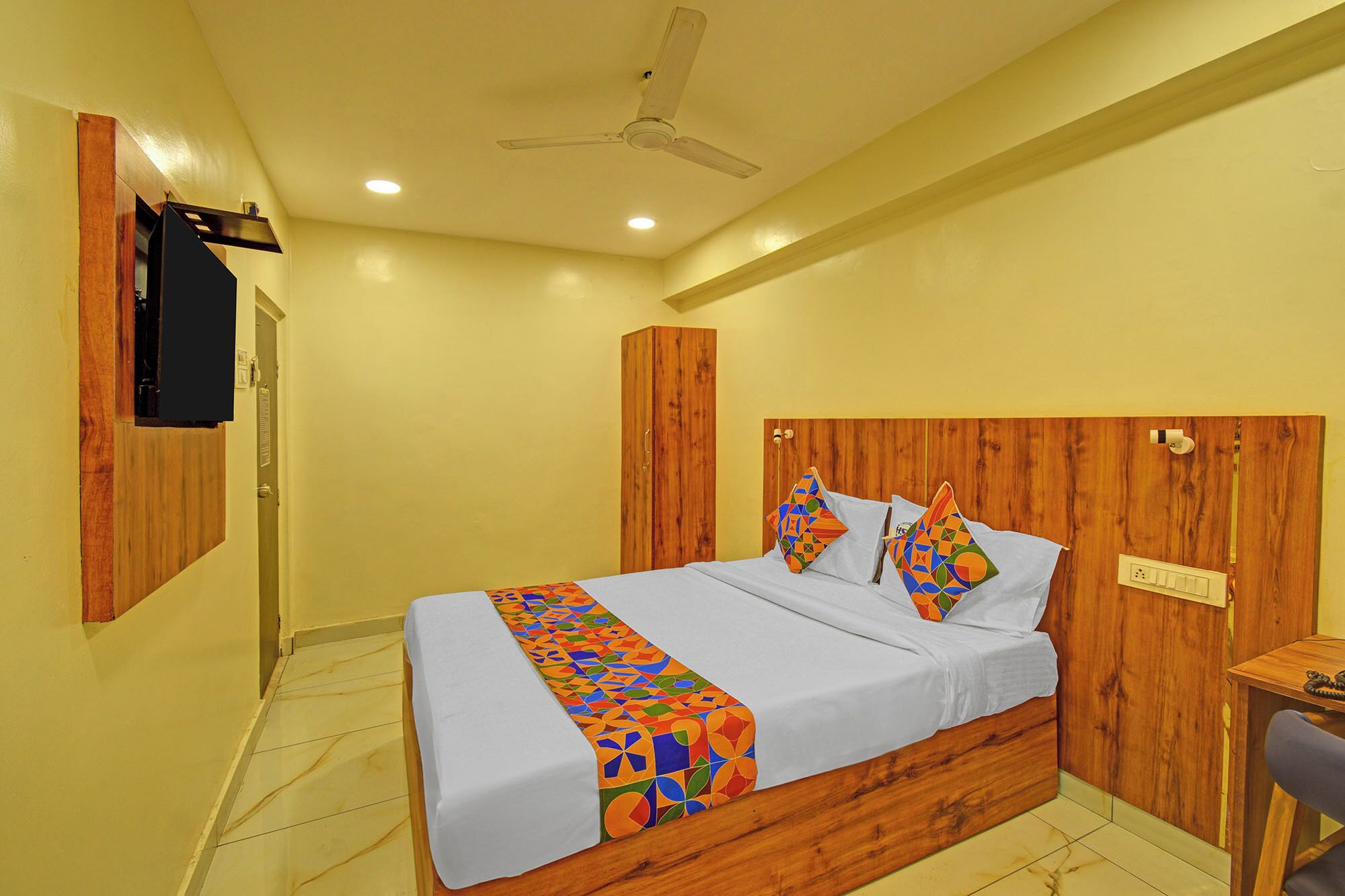 A bedroom with a wooden headboard, a bed with colorful pillows, a desk, and a ceiling fan.