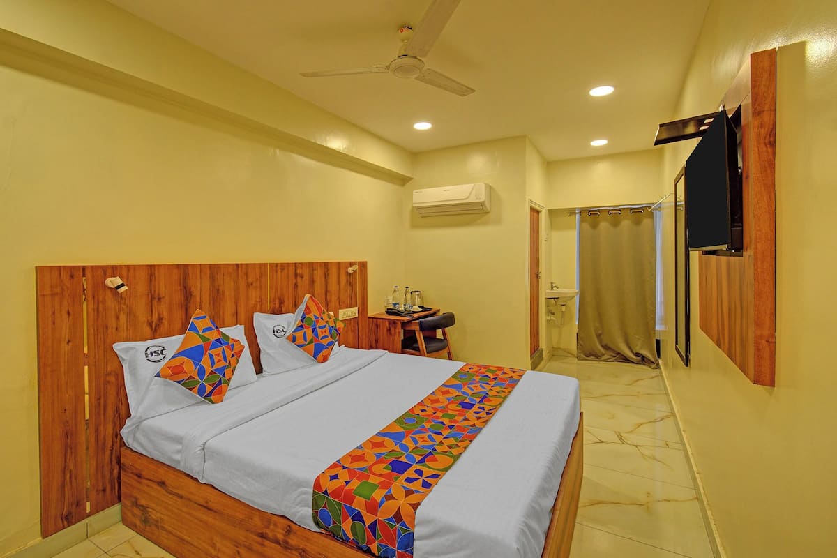 Une chambre d’hôtel avec une tête de lit en bois, un lit avec une literie colorée, un bureau avec une chaise, un appareil de climatisation, un espace douche et un téléviseur fixé au mur.