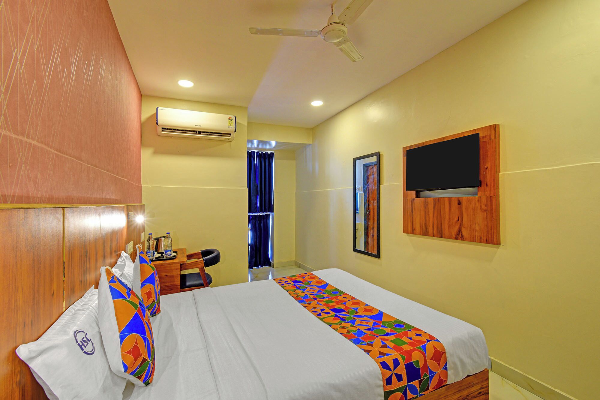 A hotel room with a bed, a desk, an air conditioner, a ceiling fan, and a TV mounted on the wall.