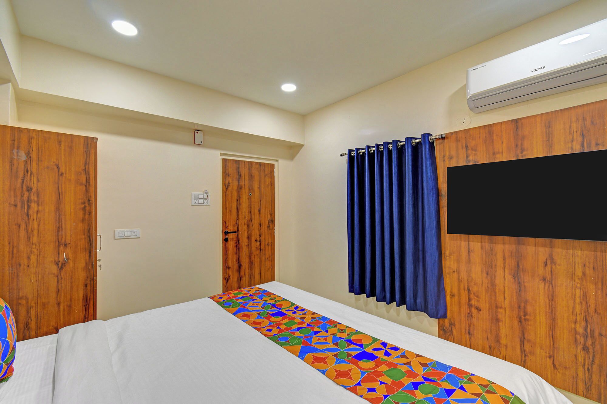 A hotel room with a bed, a wooden wardrobe, a blue curtain, an air conditioning unit, and a wall-mounted TV.