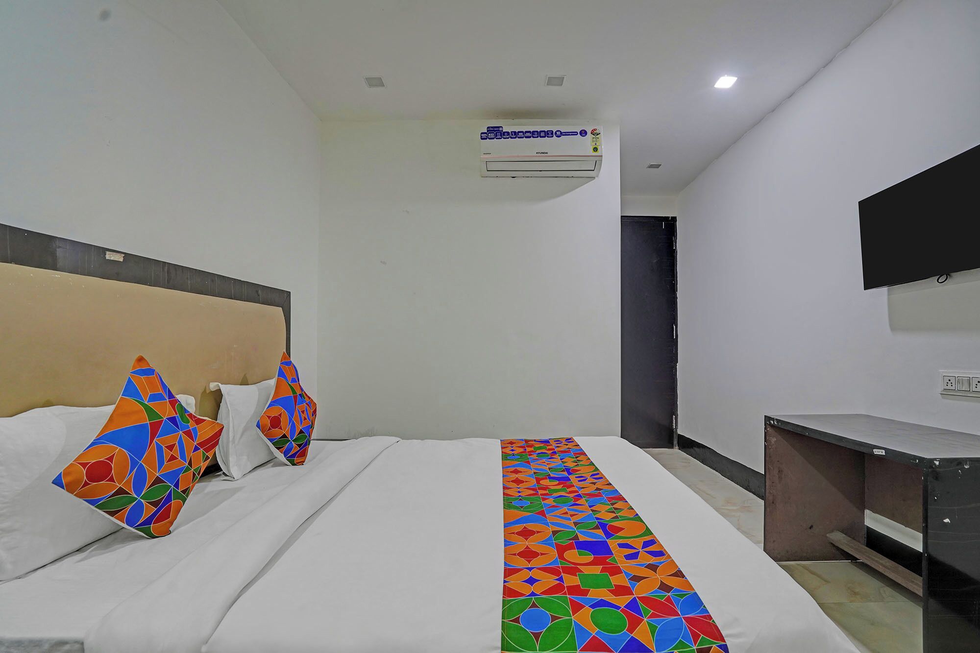 A hotel room with a bed, pillows, a desk, a flat-screen TV, and an air conditioning unit.