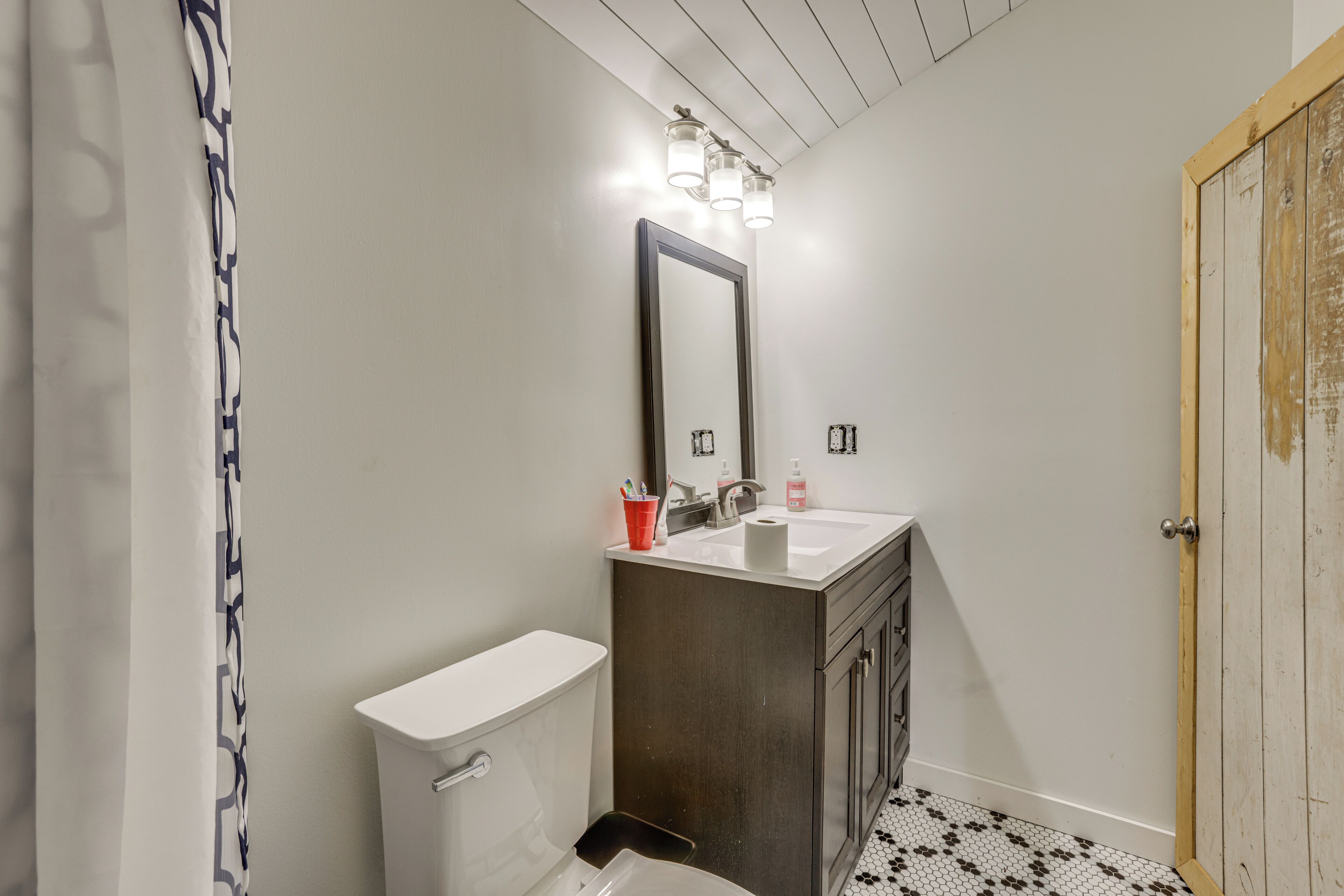 Casa de banho com sanita, lavatório e espelho. Tem cortina de chuveiro e uma porta.