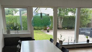 Ein Essbereich mit großem Tisch und Stühlen, großen Fenstern mit Blick auf einen Garten und einer Deckenleuchte.