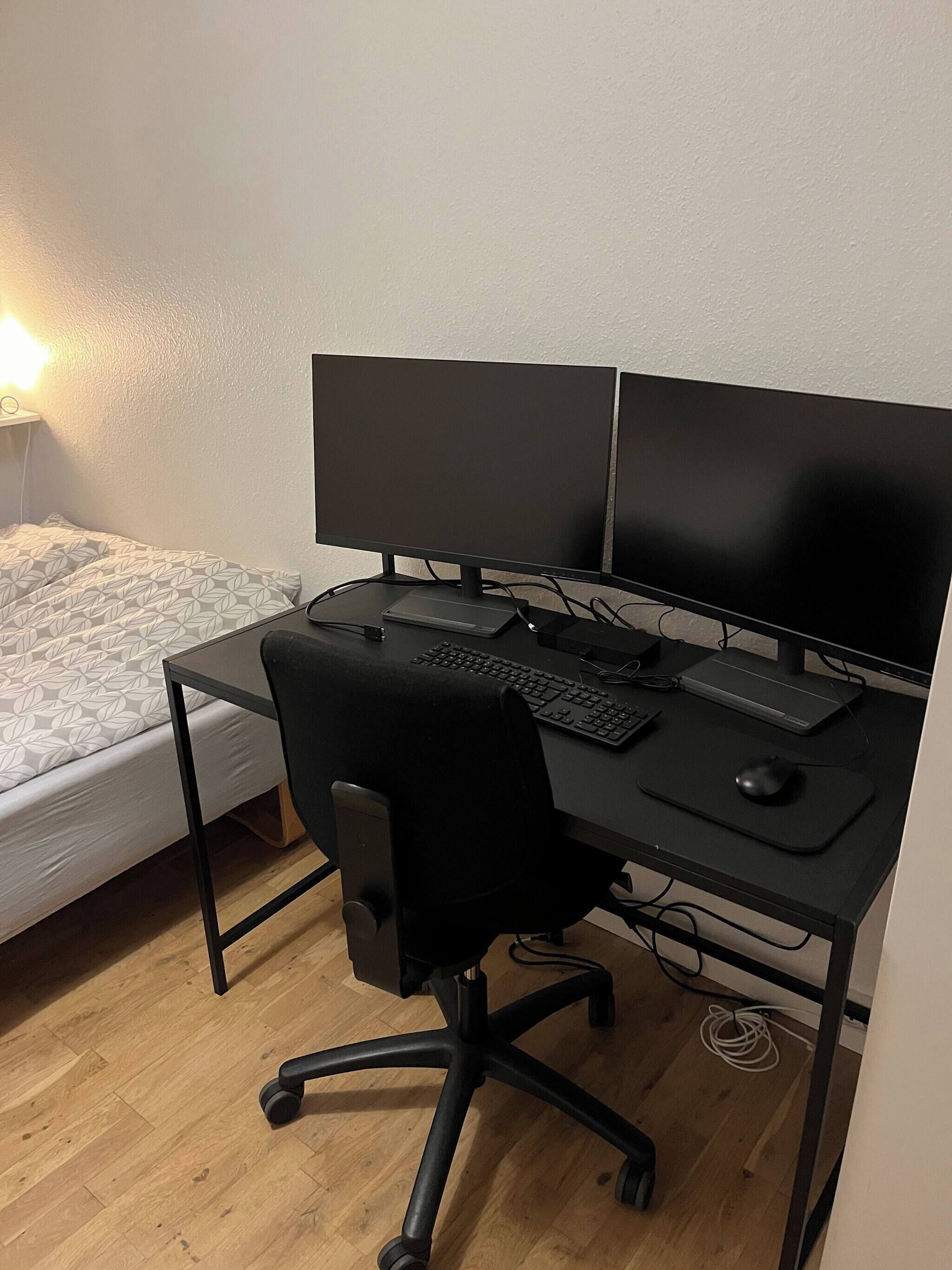 Une chambre à coucher de taille réduite, avec un lit simple, un bureau doté de deux écrans, un clavier, une souris et une chaise.