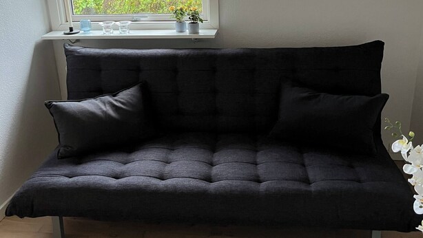 Ein dunkelgraues Sofa steht in einer Ecke unter einem Fenster.