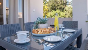 Et spisebord med morgenmadsprodukter, en potteplante og en juiceflaske på en balkon med udsigt til grønne områder.