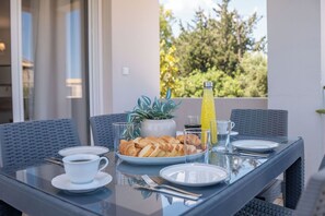 Ein Esstisch mit Frühstücksgebäck, einer Topfpflanze und einer Saftflasche auf einem Balkon mit Blick auf Grün.