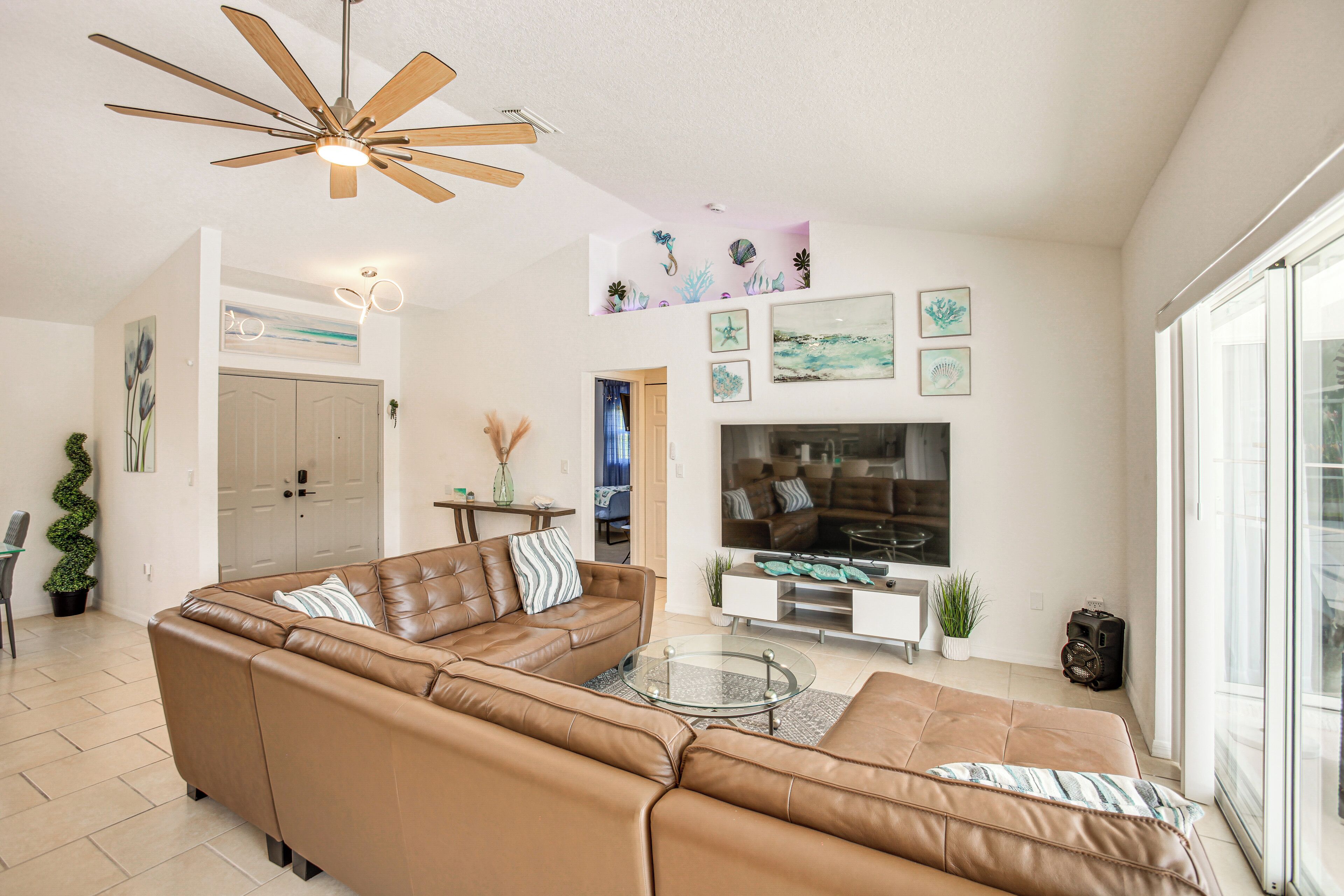 A living room with a brown leather sectional sofa, a glass coffee table, a ceiling fan, and a large flat-screen TV mounted on the wall.