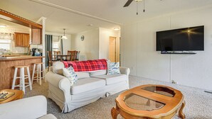 A living room with a sofa, coffee table, and a TV mounted on the wall. There is a kitchen area with wooden cabinets and a bar stool. - Steps to Lake Eufaula: Family Home w/ Deck & Yard (Eufaula)