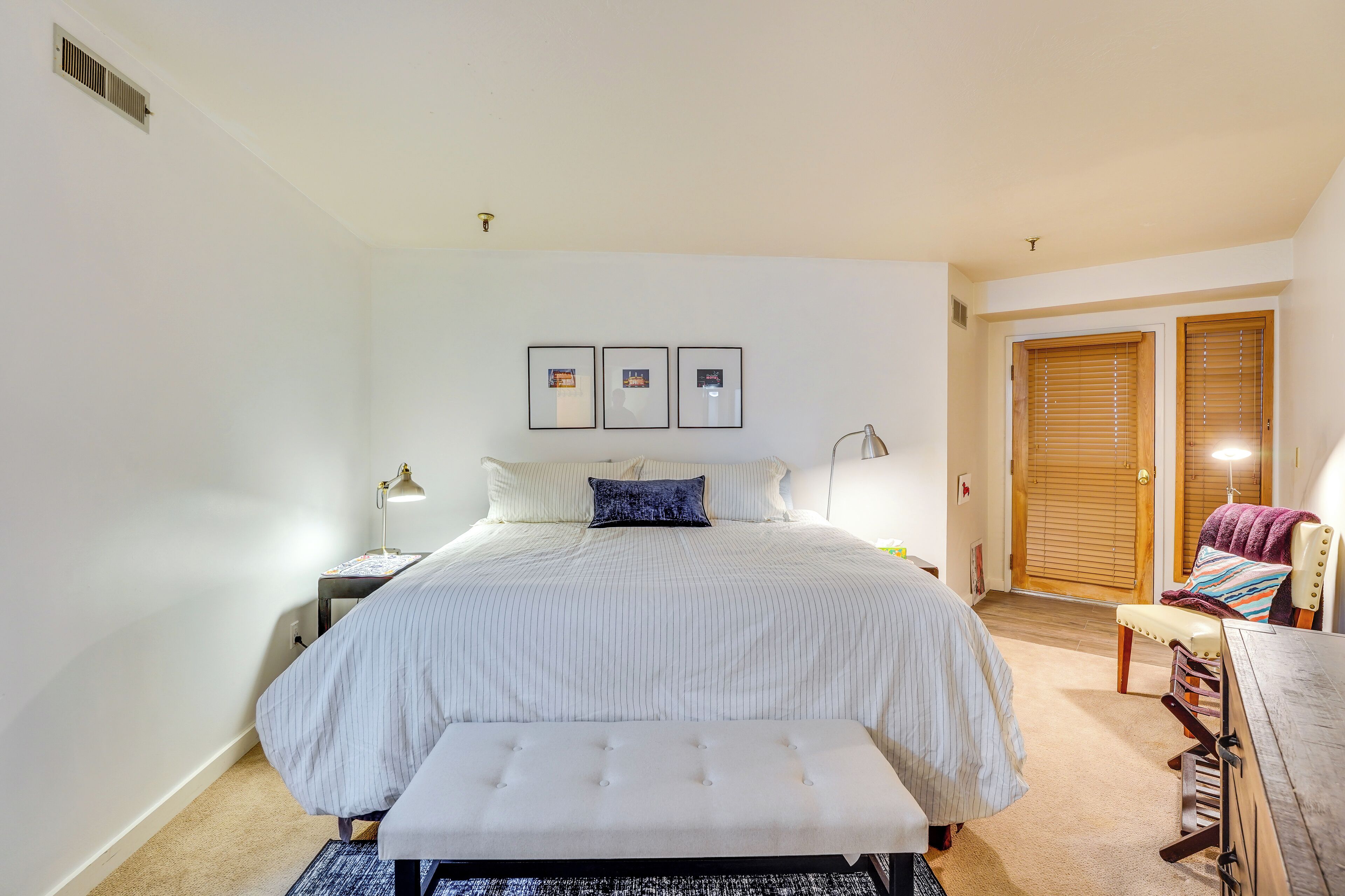 Un dormitorio con cama, mesita de noche, lámpara y un banco.