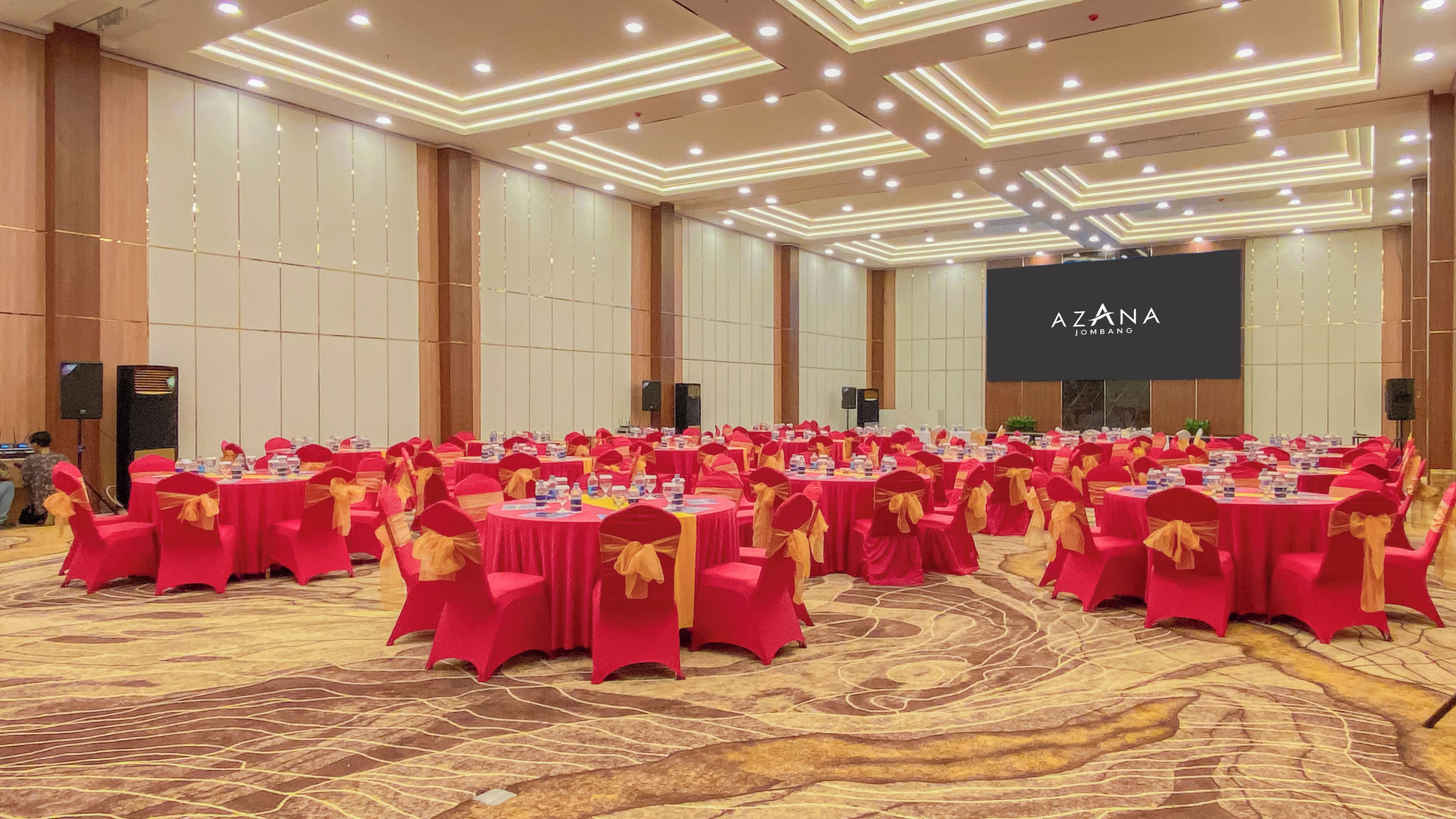 A large conference room with round tables, red and gold chairs, and a stage with a screen.