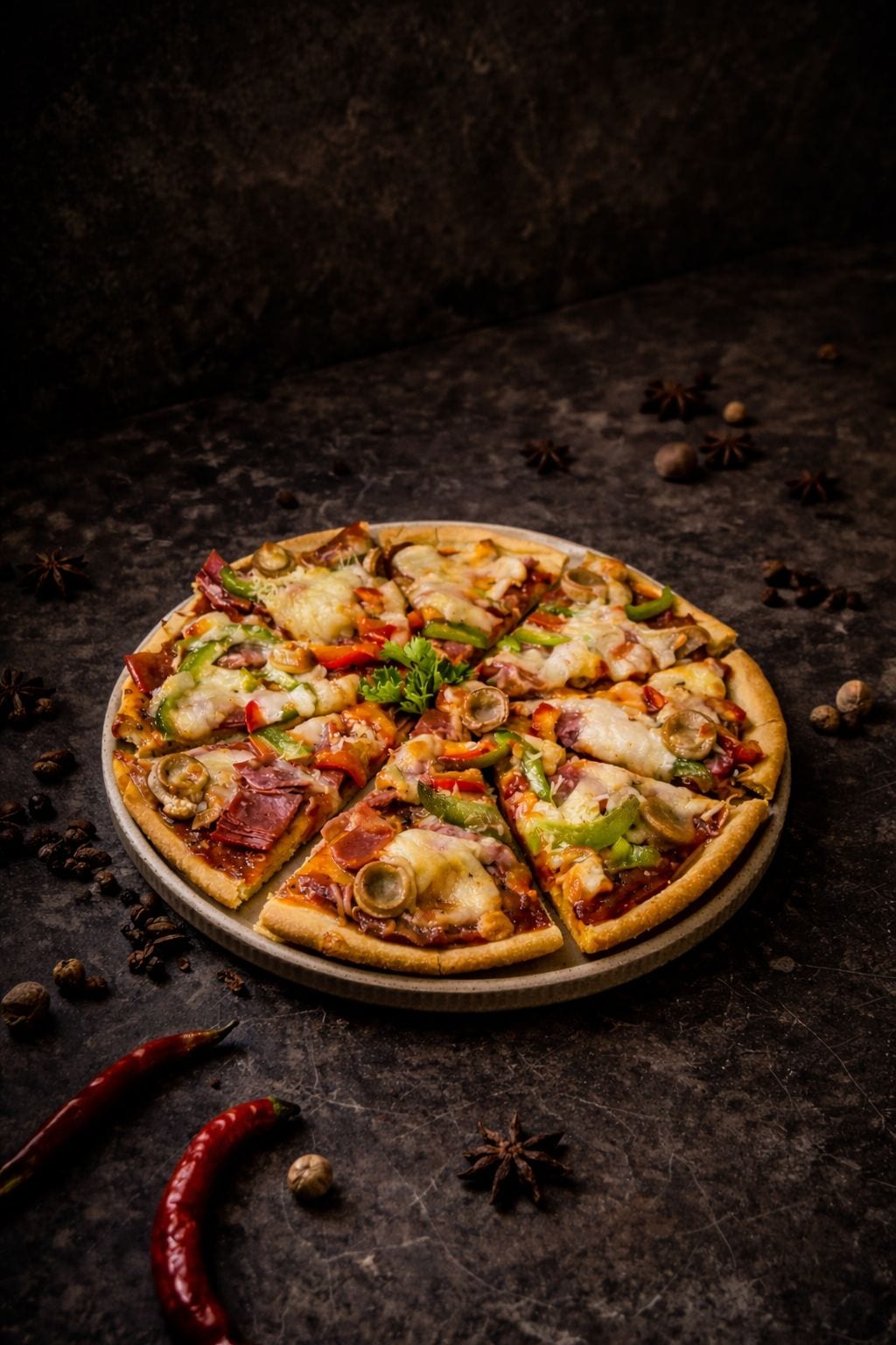 A pizza with various toppings on a round plate, surrounded by scattered spices and a dark background.