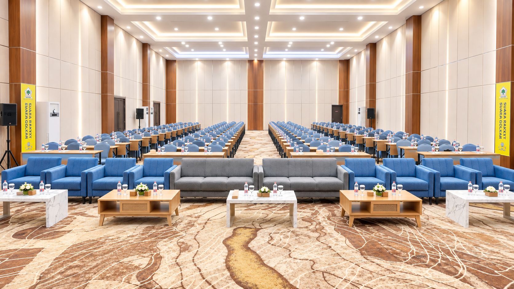 A conference room with rows of tables and chairs, a stage, and a carpeted floor.