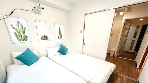 Une chambre d’hôtel avec deux lits simples, du linge de lit blanc, des coussins turquoise et des œuvres d’art sur le thème des cactus accrochées au mur.
