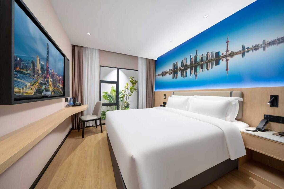 A hotel room with a large bed, a desk, a chair, a TV, and a cityscape mural on the wall.