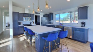 A modern kitchen with a large island, blue bar stools, and pendant lighting. - The Hills Edge - Stunning Views, Hot Tub & Games (Mariposa)