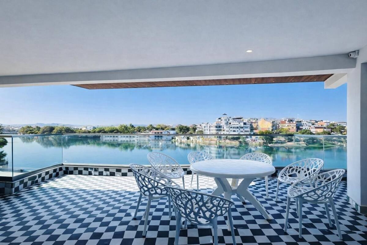 A rooftop terrace with a checkered floor, white outdoor furniture, and a view of a lake and buildings in the distance.