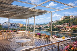 An outdoor terrace with white chairs and tables, overlooking a river and a cityscape. - Moti Mahal Lakeview Pichola Hotel (Udaipur)
