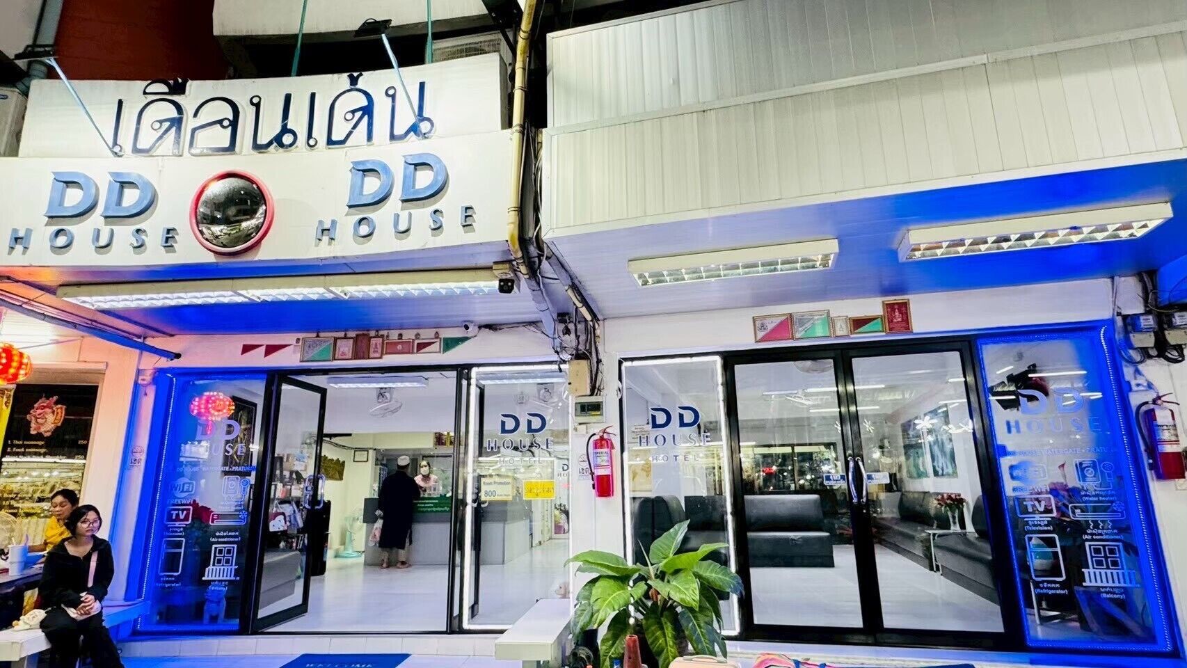 A hotel entrance with glass doors, a sign above reading "DD HOUSE HOTEL", and a blue-lit facade.