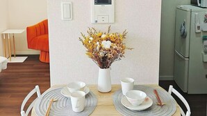Ein Essbereich mit einem Holztisch, weißen Stühlen, einer Wanduhr, einem Thermostat und einer kleinen Vase mit Trockenblumen.