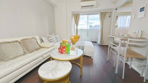 Ein Hotelzimmer mit einer weißen Couch, einem runden Tisch mit Marmorplatte, einem Bett mit weißer und goldener Bettdecke, einem kleinen weißen Tisch mit Blumenvase und einem Fenster mit Vorhängen.