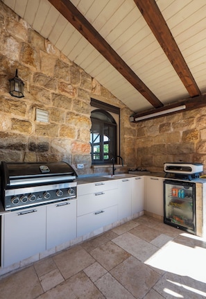 A stone-walled kitchen with a built-in grill, a refrigerator, and a window.