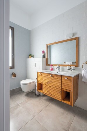 Un baño con inodoro blanco, tocador de madera con espejo y lavamanos blanco.