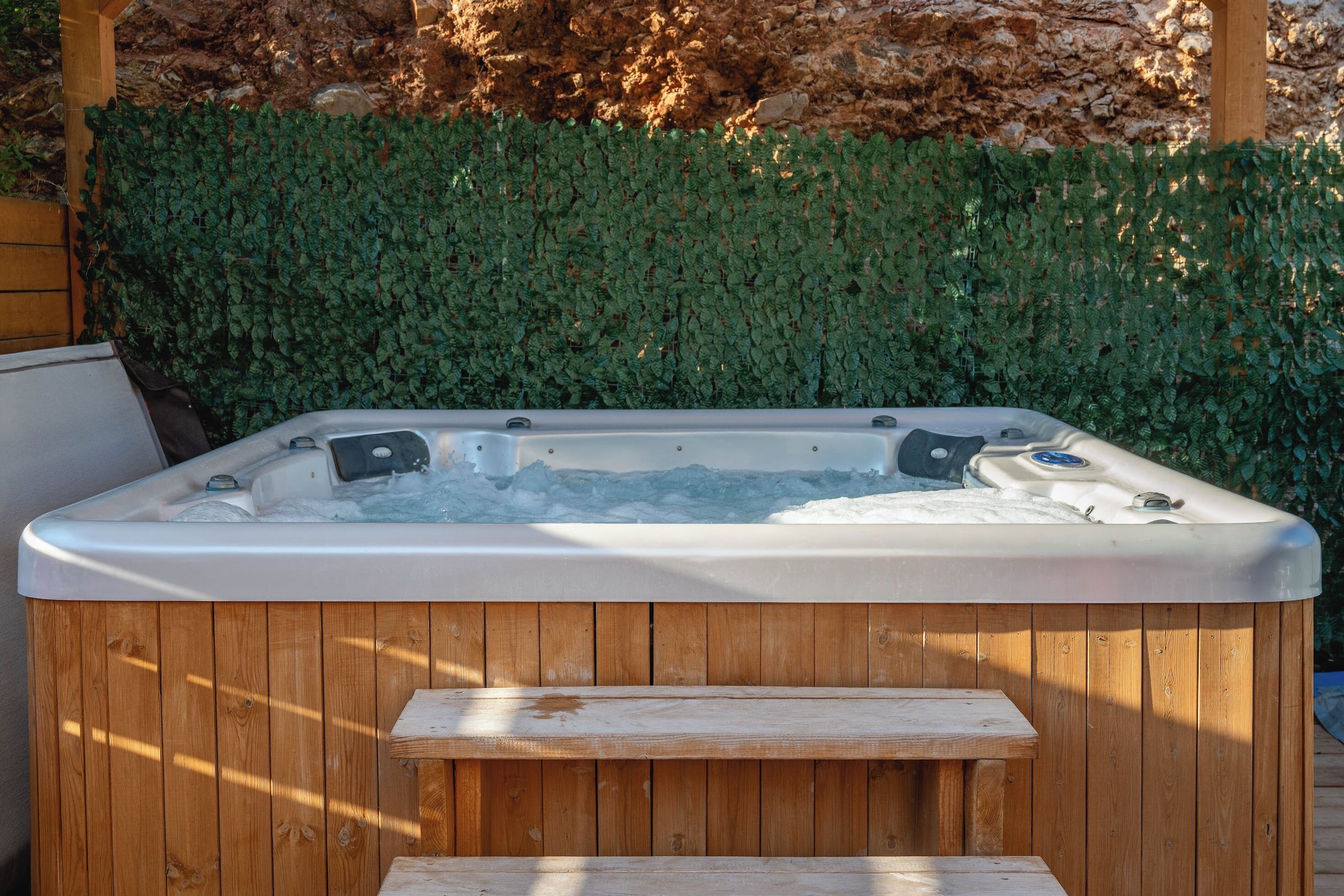 A wooden hot tub with a bench in front, surrounded by a wooden deck and a green hedge.