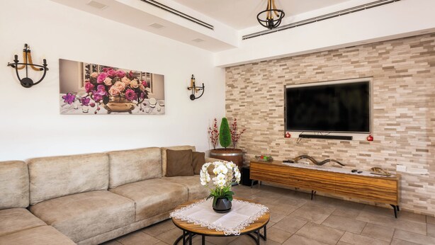 Un salón con un sofá beige, una mesa de centro redonda con un florero, un televisor de pantalla plana montado en una pared con textura y una consola de madera.