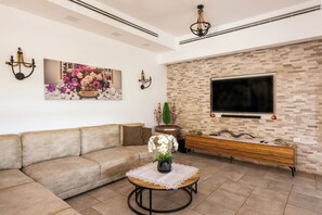Un soggiorno con un divano beige, un tavolino rotondo con un vaso di fiori, un televisore a schermo piatto montato su una parete strutturata e un tavolino consolle in legno.