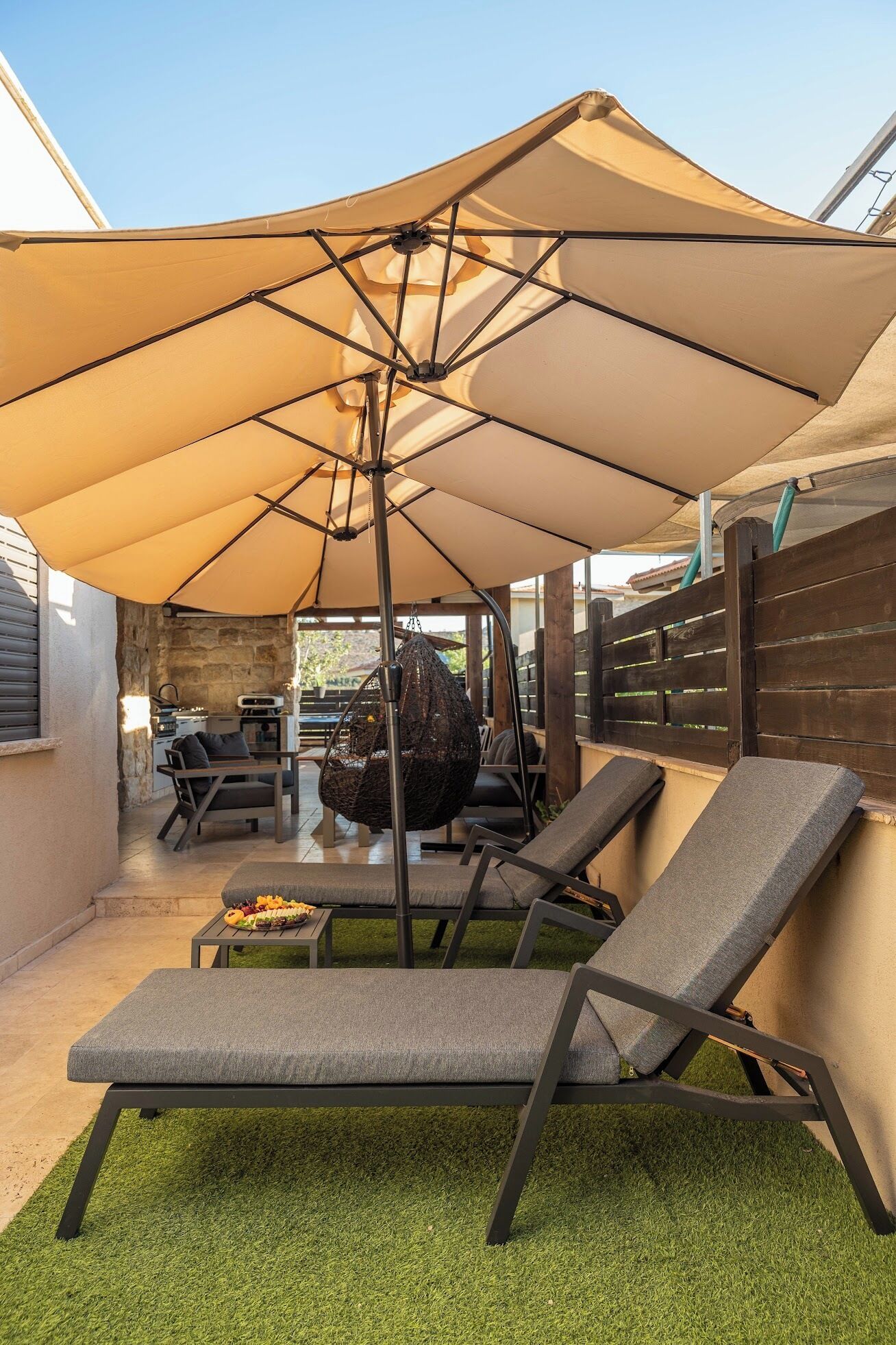 Ein Terrassenbereich im Freien mit einem großen Sonnenschirm, zwei Liegestühlen, einem Hängesessel und einem kleinen Tisch mit Speisen.