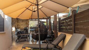 Ein Terrassenbereich im Freien mit einem großen Sonnenschirm, zwei Liegestühlen, einem Hängesessel und einem kleinen Tisch mit Speisen.