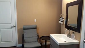 A bathroom with a white door, a grey chair, a small round table, a white sink with a mirror, and a dark cabinet. - Small, renovated, private 1 bedroom bathroom in front of bus stop to downtown. (Vancouver)