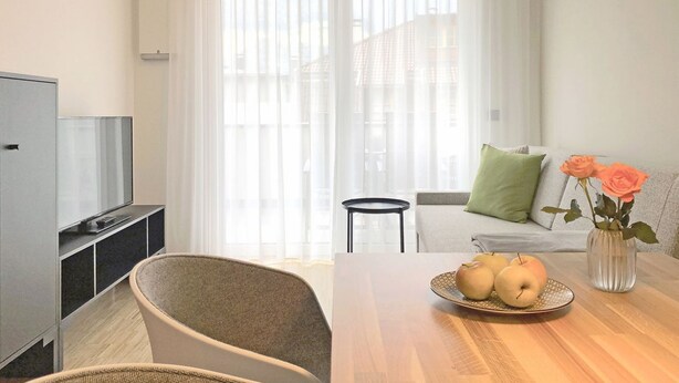 Una sala de estar moderna con una televisión, una mesa de comedor con manzanas y un jarrón de flores, un sofá con cojines y una silla.