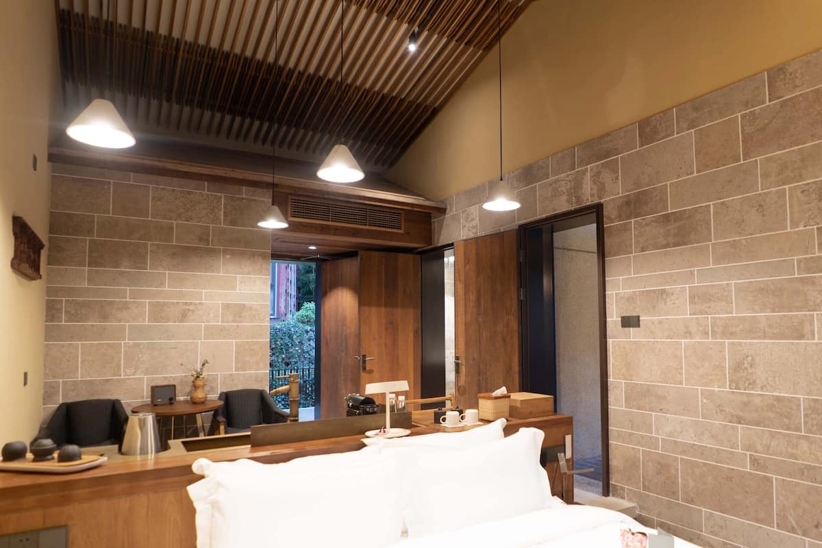 A modern hotel room with a large bed, wooden ceiling, and stone walls.