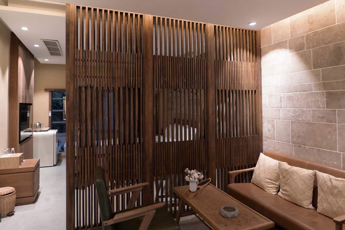 A modern hotel room with a wooden partition, a sofa, a small table, and a bathroom visible in the background.