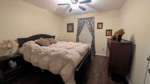 Un dormitorio con cama, mesita de noche, cómoda, ventilador de techo y ventana con cortinas.