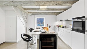 A modern kitchen with white cabinetry, a built-in oven, and a dining area with a bar stool. - Casa la Tortue 2 Scaiano for 6 (Gambarogno)