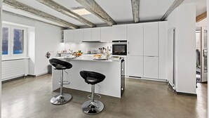 A modern kitchen with white cabinetry, a central island with bar stools, exposed wooden beams, and a view of the outdoors through a window. - Casa la Tortue 2 Scaiano for 6 (Gambarogno)