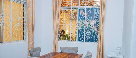 A dining area with a wooden table and grey chairs, a ceiling fan, decorative window grills, and a sink.