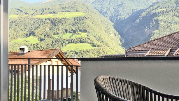 A balcony with a view of a mountainous landscape, a wooden chair, and a building with a tiled roof. - Apartment Luis 2 - Alpstay (Naturns)