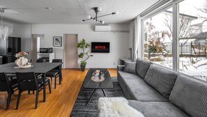 Una sala de estar moderna con comedor, ventanas amplias y un televisor de pantalla plana mostrando 'NETFLIX'.