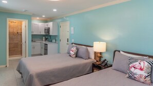 A bedroom with two beds, a nightstand with a lamp, a phone, and a kitchenette in the background. - Flamingo Inn - 2 Queen with Kitchenette #204 (Fort Myers Beach)