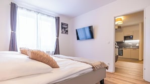 Un dormitorio con cama, cortinas, televisor de pantalla plana y vista a una cocina.