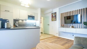Una cocina moderna con armarios blancos, microondas integrado y cafetera. En la zona de estar, hay una mesa de comedor de madera y un sofá gris. Un televisor de pantalla plana está montado en la pared sobre el sofá.