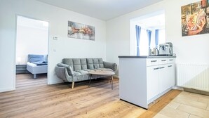 Una sala moderna con un sofá gris, una mesita de centro redonda y una cocina con encimera y armarios blancos.