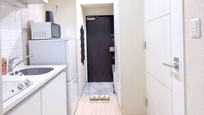 Un couloir étroit avec un coin cuisine sur la gauche, comprenant un four à micro-ondes, un évier et des placards. On peut voir trois paires de chaussures sur le sol, et une porte au fond du couloir.