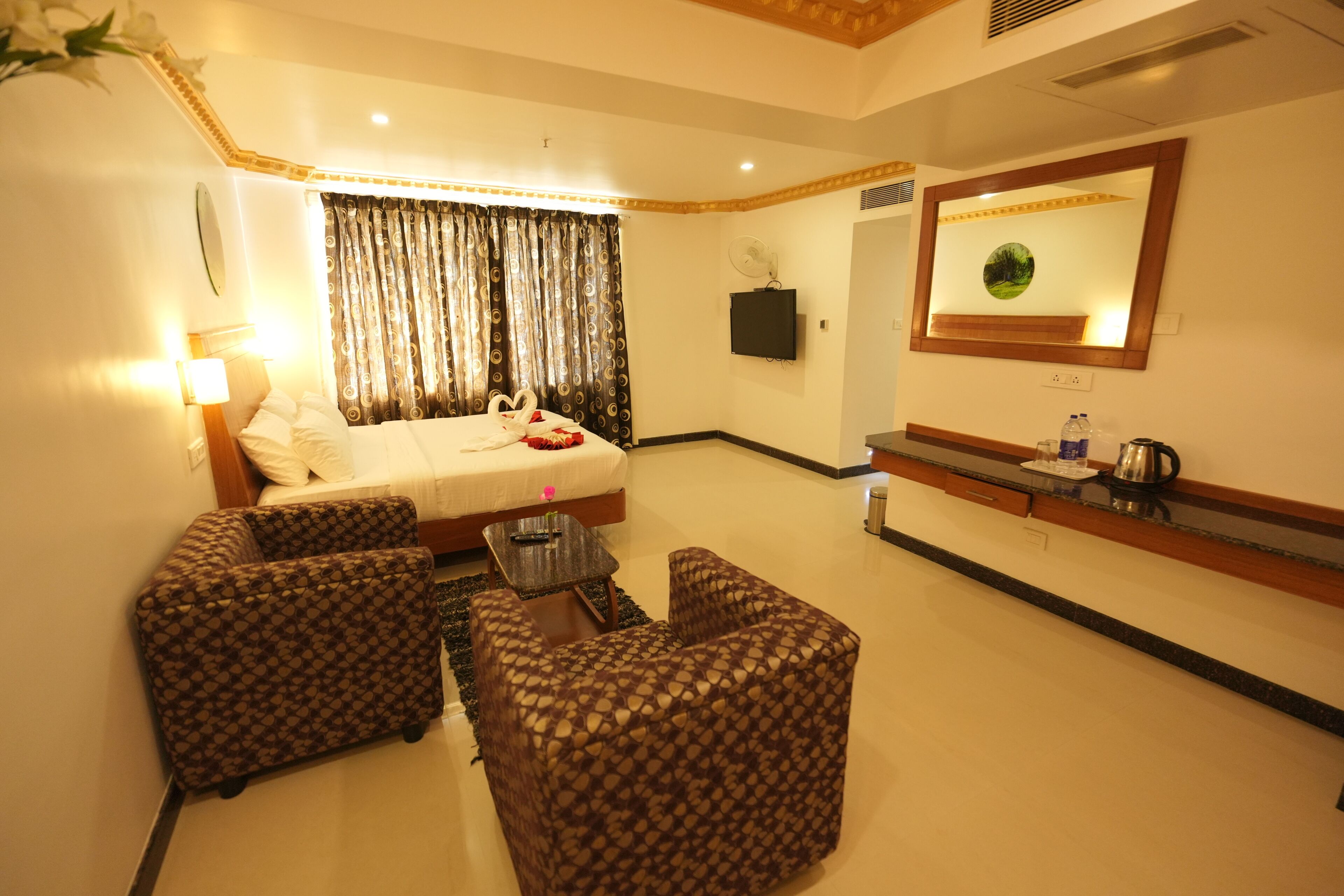 A hotel room with a bed, two patterned armchairs, a small table, a TV, a mirror, and a coffee maker.