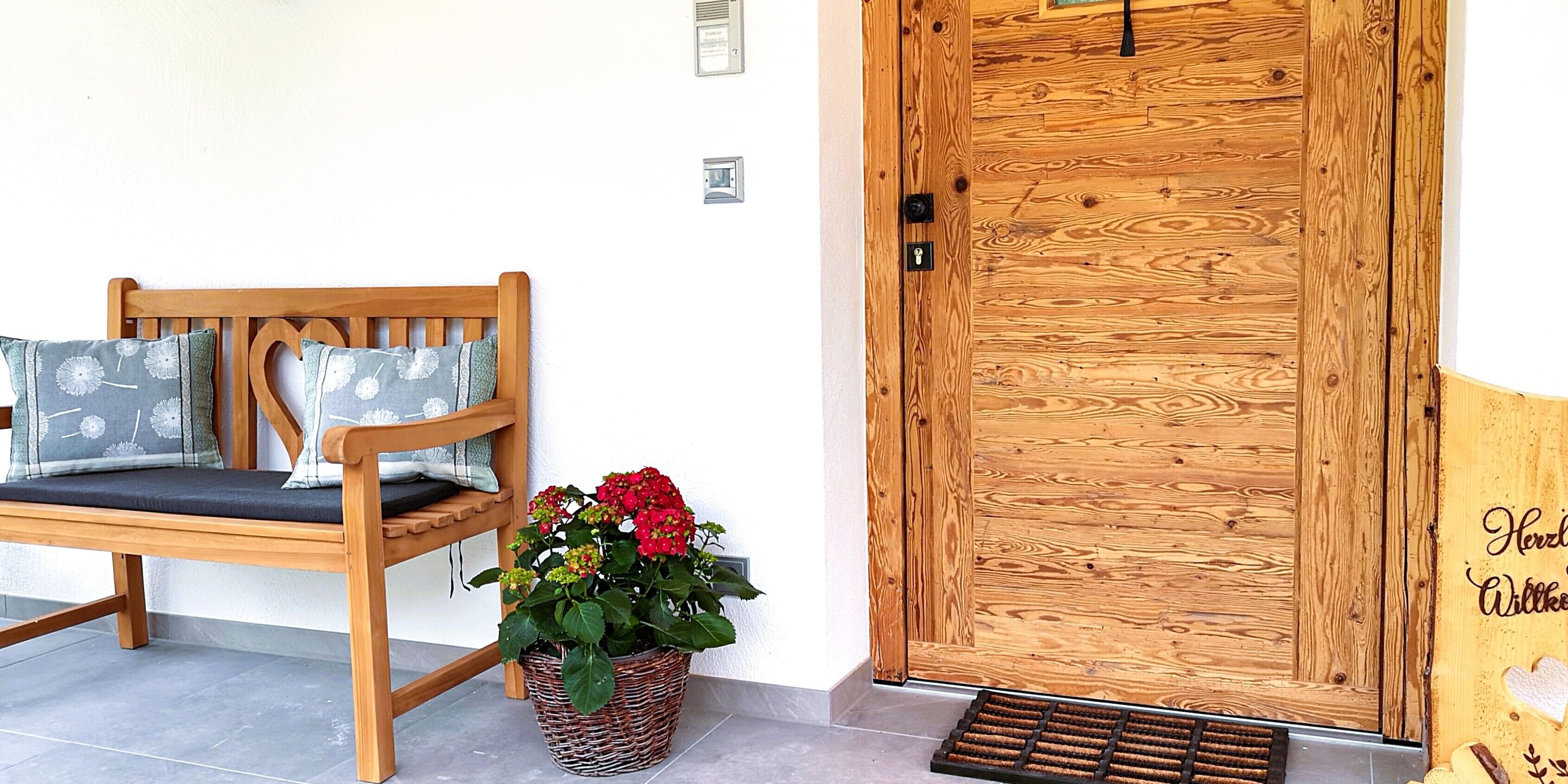 A wooden bench with cushions, a wicker plant pot with flowers, and a wooden door with a welcome sign.